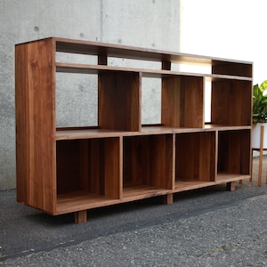 May include: A wooden bookshelf with multiple open compartments and shelves. The bookshelf is made of dark brown wood and has a modern design. It is placed against a concrete wall and a white wall. A potted plant is next to the bookshelf.