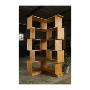 May include: A light-colored wooden bookshelf with a unique, geometric design. The bookshelf features multiple open cubby spaces of varying sizes, arranged in a staggered, angular configuration. The wood has a natural finish, showcasing the grain.