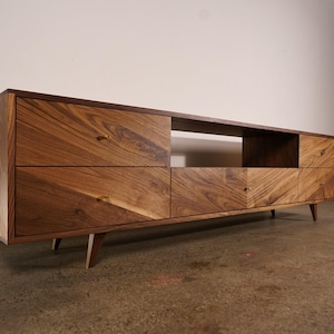 May include: A mid-century modern style wooden media console with four drawers and an open shelf. The console is made of a warm-toned wood with a chevron pattern on the drawer fronts. It has angled legs and brass hardware.