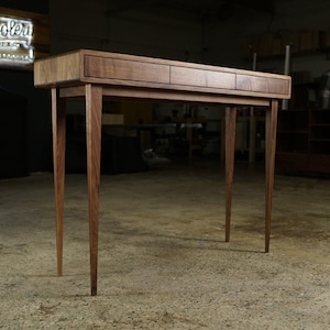 May include: A handcrafted wooden console table with three drawers and tapered legs. The table is made of dark wood and has a natural finish. The design is minimalist and modern, suitable for a living room or entryway.