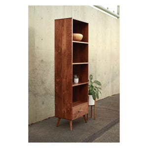 May include: A tall, mid-century modern wooden bookshelf with four shelves and a drawer. The bookshelf is made of dark wood and has angled legs. A small plant and a wooden bowl are visible on the shelves. The bookshelf is approximately 183 cm tall.