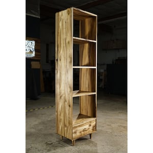 May include: A tall wooden bookcase with multiple shelves and a drawer. The bookcase is made of light-coloured wood with visible grain patterns. It has a rectangular shape and stands on four short legs. The design is simple and modern, suitable for storage and display.