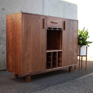 May include: A wooden bar cabinet with a warm brown finish. It features two doors, a drawer, and an open section for wine glasses and bottles. The cabinet is raised on short legs and has a minimalist design. A potted plant is next to the cabinet.