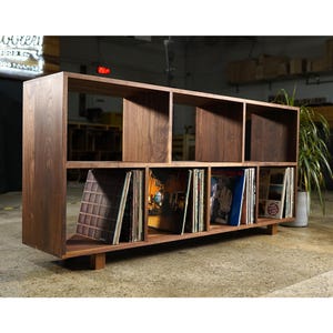 May include: A dark brown wooden record storage unit with multiple compartments. The unit is rectangular with a low profile and is filled with vinyl records. The unit is elevated on short wooden legs.