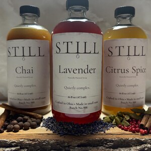 May include: Three glass bottles of flavored syrup with black caps, labeled "Chai", "Lavender", and "Citrus Spice". Each bottle is 16 fl oz (473 ml). Spices and herbs are arranged in front of the bottles on a wooden board.
