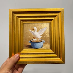 Puede incluir: Pintura enmarcada que representa un pato blanco con las alas extendidas en una olla azul. La obra de arte tiene un fondo dorado y está enmarcada en un marco dorado texturizado. La pintura está sujeta por una mano.