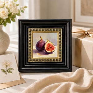 May include: A framed painting of two figs, one whole and one sliced, showcasing a vibrant interior. The artwork is set in a black frame with a gold inner border. The painting is on a white surface.