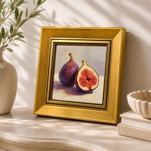 May include: A framed painting of two figs, one whole and one halved, with a gold frame. The figs are purple with red interiors, set against a neutral background. The artwork is displayed on a white surface.