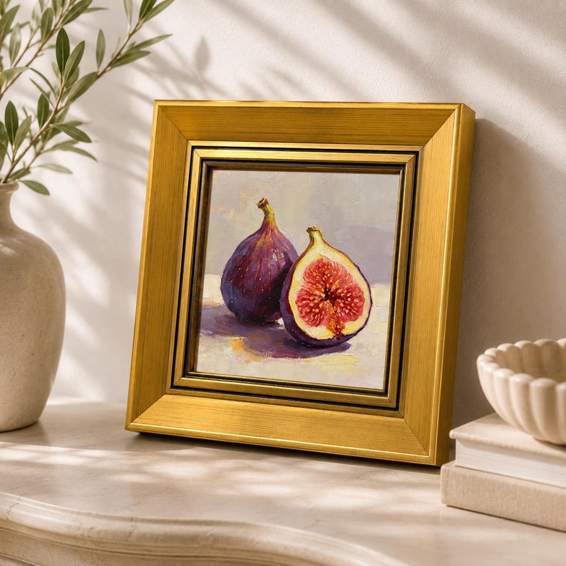 May include: A framed painting of two figs, one whole and one halved, with a gold frame. The figs are purple with red interiors, set against a neutral background. The artwork is displayed on a white surface.