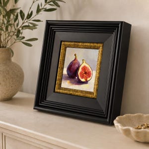 May include: A framed painting of two figs, one whole and one cut open, revealing the red interior. The painting is set in a black frame with a gold inner border. The artwork is displayed on a white surface.