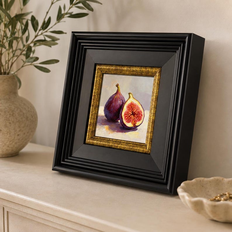 May include: A framed painting of two figs, one whole and one cut open, revealing the red interior. The painting is set in a black frame with a gold inner border. The artwork is displayed on a white surface.
