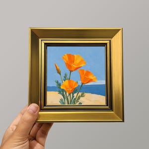 以下が含まれることがあります： メタリックゴールドのフレームに入った正方形の絵画。明るい青空と砂浜の風景を背景に、鮮やかなオレンジ色のカリフォルニアポピーが3輪描かれており、背景には濃い青色の海の水平線が見えるアート作品。