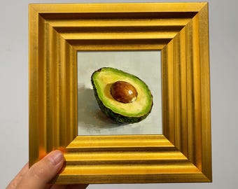 May include: A framed oil painting of a halved avocado. The avocado is green with a brown pit, set against a light background. The painting is in a gold-colored frame.