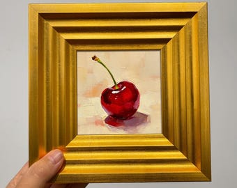 May include: A small, square painting of a single, ripe cherry with a green stem. The cherry is a deep red color and is set against a light background. The painting is framed in a gold, textured frame.