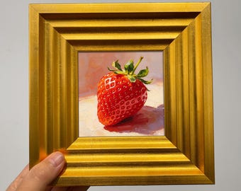 May include: A small square oil painting of a single red strawberry with green leaves, rendered in a realistic style. The artwork is encased in a thick, ornate, metallic gold frame with stepped, beveled edges and a polished, reflective finish.