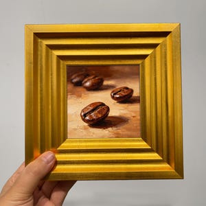 May include: A small, square painting of coffee beans in a gold frame. The artwork features several brown coffee beans on a light brown background. The frame is a bright gold color with a textured design.