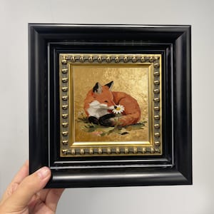 Könnte beinhalten: Gerahmtes Gemälde eines Fuchses, der auf Gras ruht und eine Gänseblümchen hält. Der Fuchs hat orange-weißes Fell mit schwarzer Nase und Ohren. Das Gemälde befindet sich in einem goldenen Rahmen mit einem schwarzen Außenrahmen.