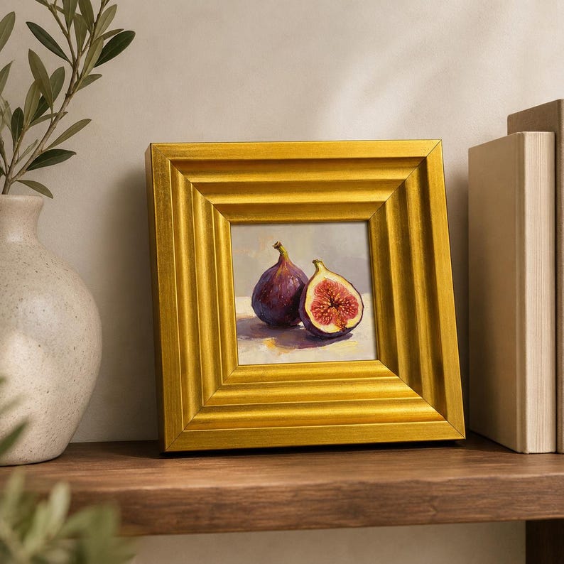 May include: A square, gold-framed painting of two figs, one whole and one cut open, revealing the red interior. The painting is on a wooden shelf, with a vase and books in the background.