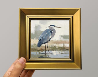 May include: A square painting of a blue heron standing in shallow water, rendered in a painterly style with muted tones of blue, grey, and tan. The artwork is encased in a thick, ornate, metallic gold frame with a stepped, beveled profile.