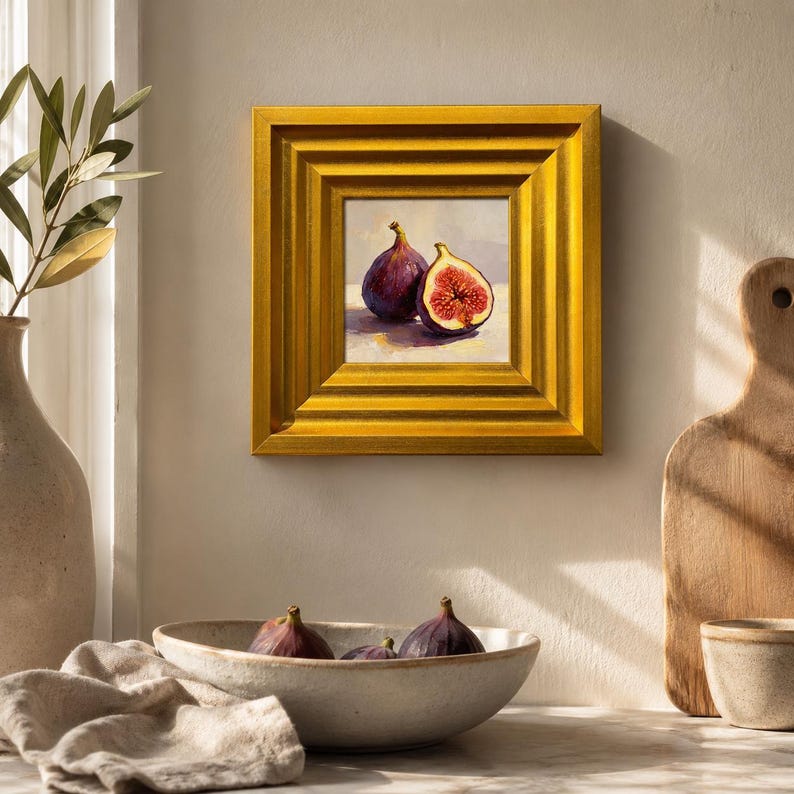 May include: A framed painting of two figs, one whole and one cut open, with a golden frame. The painting is displayed on a neutral wall. A ceramic bowl with figs and a cutting board are in the foreground.
