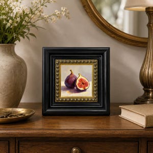 May include: A framed painting of two figs, one whole and one sliced, showcasing a rich purple exterior and vibrant red interior. The artwork is set in a black frame with a gold inner border, displayed on a wooden surface.