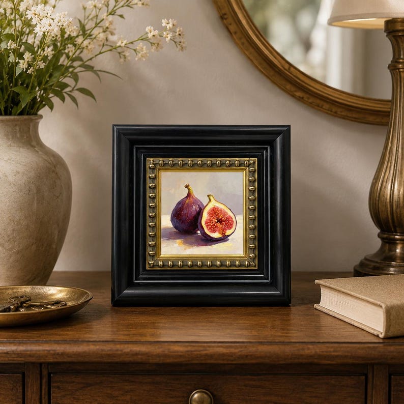 May include: A framed painting of two figs, one whole and one sliced, showcasing a rich purple exterior and vibrant red interior. The artwork is set in a black frame with a gold inner border, displayed on a wooden surface.