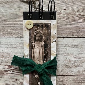 May include: A rectangular spiral-bound notebook featuring a vintage-style black and white photograph of a young person in a dress, accented with a cream-colored button, a dark green fabric ribbon tied in a bow, and a silver metal lobster clasp.