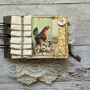 May include: A spiral-bound journal featuring a central illustration of a hen and chicks. The cover includes cream-colored fabric ruffles, a white button, a mustard yellow patterned section, and a small metal key attached to the side.
