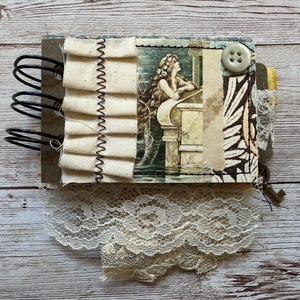May include: A spiral-bound journal featuring a collage cover with a mermaid illustration, cream-colored fabric ruffles with black stitching, a white button, and a black and white patterned paper section, finished with a layer of white lace trim.