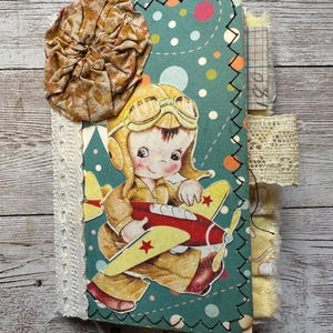 May include: A handmade journal with a vintage aesthetic. The cover features a teal background with a cartoon pilot and airplane. Embellishments include a fabric flower, lace trim, and decorative stitching. The journal is tied with a ribbon.