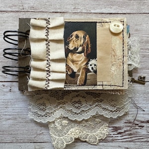 May include: A spiral-bound journal featuring a vintage-style illustration of a brown dog and a small white dog. The cover is decorated with a cream-colored fabric ruffle, a white button, and layers of white lace trim attached to the bottom edge.