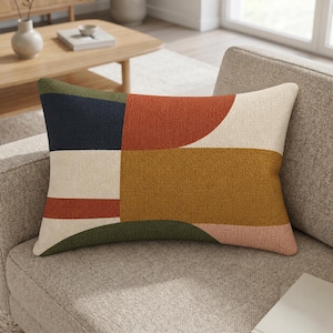 Könnte beinhalten: Rechteckiges Zierkissen mit geometrischem Muster. Das Kissen hat einen strukturierten Stoff mit Farbblöcken in Marineblau, Rostrot, Olivgrün, Senfgelb, Creme und Rosa. Es liegt auf einem grauen Sofa.