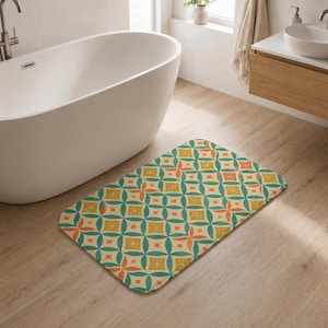 May include: A rectangular bath mat featuring a repeating geometric pattern of teal circles, orange four-pointed stars, and mustard yellow squares with small white starburst accents. The mat has rounded corners and a soft, flat-weave fabric texture.