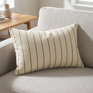 May include: A rectangular throw pillow featuring a cream-colored fabric base with vertical olive green stripes. The pillow has a textured appearance and is positioned on a light gray upholstered sofa, showcasing a clean and simple design.