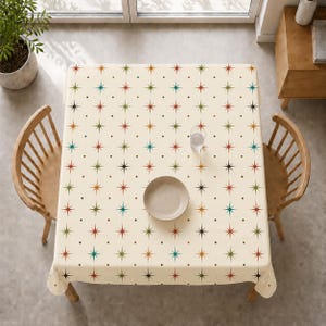 May include: A cream-coloured square tablecloth featuring a repeating pattern of colourful starbursts and small dots. A plate and a glass of water sit on the table. Two wooden chairs are positioned on either side of the table.