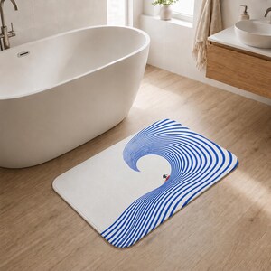 May include: A rectangular bath mat featuring a white background with a large, stylized blue wave graphic composed of concentric curved lines. A small figure on a red surfboard is positioned at the base of the wave, creating a minimalist visual effect.