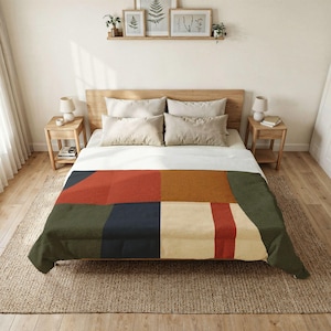 Pode incluir: Uma manta retangular com um padrão geométrico de blocos de cor em tons de terracota, amarelo mostarda, verde oliva escuro, azul marinho e creme. A manta está estendida sobre uma cama com almofadas brancas e uma cabeceira de madeira.