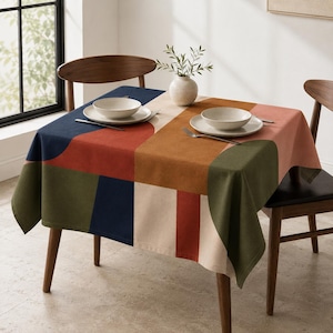 Könnte beinhalten: Eine quadratische Tischdecke mit einem geometrischen Farbblockmuster in Marineblau, Olivgrün, Terrakotta und Creme. Der Tisch ist mit zwei weißen Keramikschalen, zwei silbernen Gabeln und einer kleinen weißen Vase mit grünen Zweigen gedeckt.