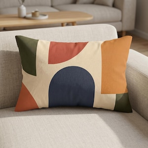 May include: Rectangular decorative pillow with a cream background and abstract shapes in rust, navy, olive green, and orange. The pillow is resting on a light gray sofa.