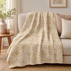 May include: A cream-colored throw blanket with a repeating fan-shaped pattern in gold. The blanket is draped over a beige sofa, with a neutral-toned pillow visible. The blanket's design adds a touch of elegance to the room.