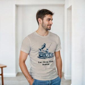 Puede incluir: Camiseta beige claro con un diseño de pesca: un pescador en un bote y un pez saltando. El texto "Eat, Sleep, Fish, Repeat" está impreso debajo. La camiseta la lleva una persona.