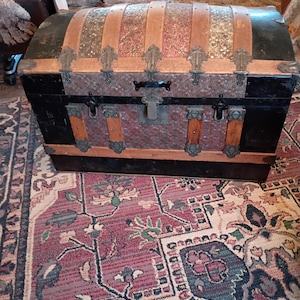 May include: Antique trunk with a domed top. The trunk features black sides, decorative metal accents, and wooden slats with patterned fabric. The trunk has a metal lock and hinges. The trunk is sitting on a patterned rug.