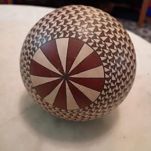 May include: A round, decorative ceramic object with a geometric pattern. The design features a central sunburst pattern in red and cream, surrounded by a series of triangles in a repeating pattern. The overall color scheme is earthy and warm.