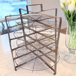 May include: An antique metal wire rack with multiple circular shelves. The rack has a rectangular frame and a handle at the top. The metal has a rustic, aged appearance. The rack is designed to hold items and is suitable for kitchen or home decor.