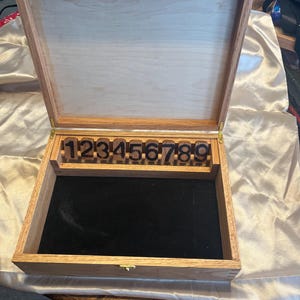 May include: Open wooden box with a light-colored interior and a dark velvet base. The box contains wooden numbers 1 through 9. The box is made of light brown wood with brass hinges and a clasp.