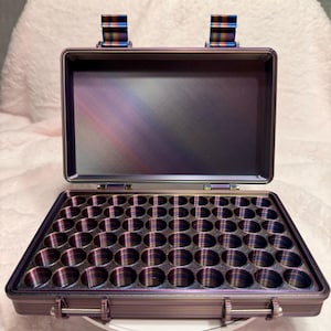 May include: An open, iridescent purple case with a grid of circular compartments. The case has a hinged lid and two multi-colored accent pieces on top. The case is designed to hold small items, and is resting on a white surface.