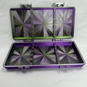 May include: A rectangular, open storage case featuring a vibrant purple and green geometric starburst pattern on the interior base and lid. The case includes two silver-colored metal latches at the top and internal dividers with small notches.