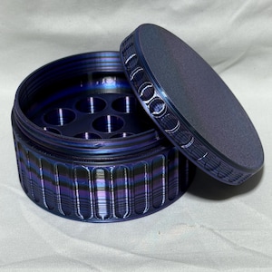 May include: A round, blue metal container with a lid. The container has a series of holes inside and a ridged exterior. The lid is slightly ajar, revealing the interior. The container has a metallic sheen, with hints of purple and green.