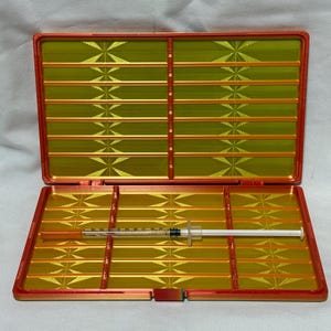 May include: Open orange and gold case with a syringe inside. The case has a rectangular shape with a gold interior and orange trim. The syringe is clear with a white plunger and needle.
