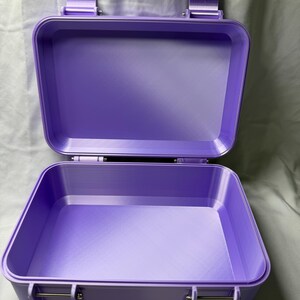 May include: Open, lavender-colored, rectangular case with a hinged lid and metal clasps. The case is made of a solid material and has a smooth, matte finish. The interior is empty, revealing the case's depth and structure. The case is designed for storage.